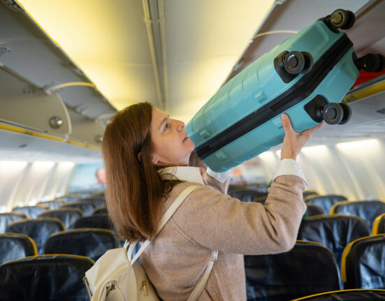 Side view of young female in casual outfit with backpack lifting luggage into compartment of airplane cabin while standing by leather chairs inside airplane