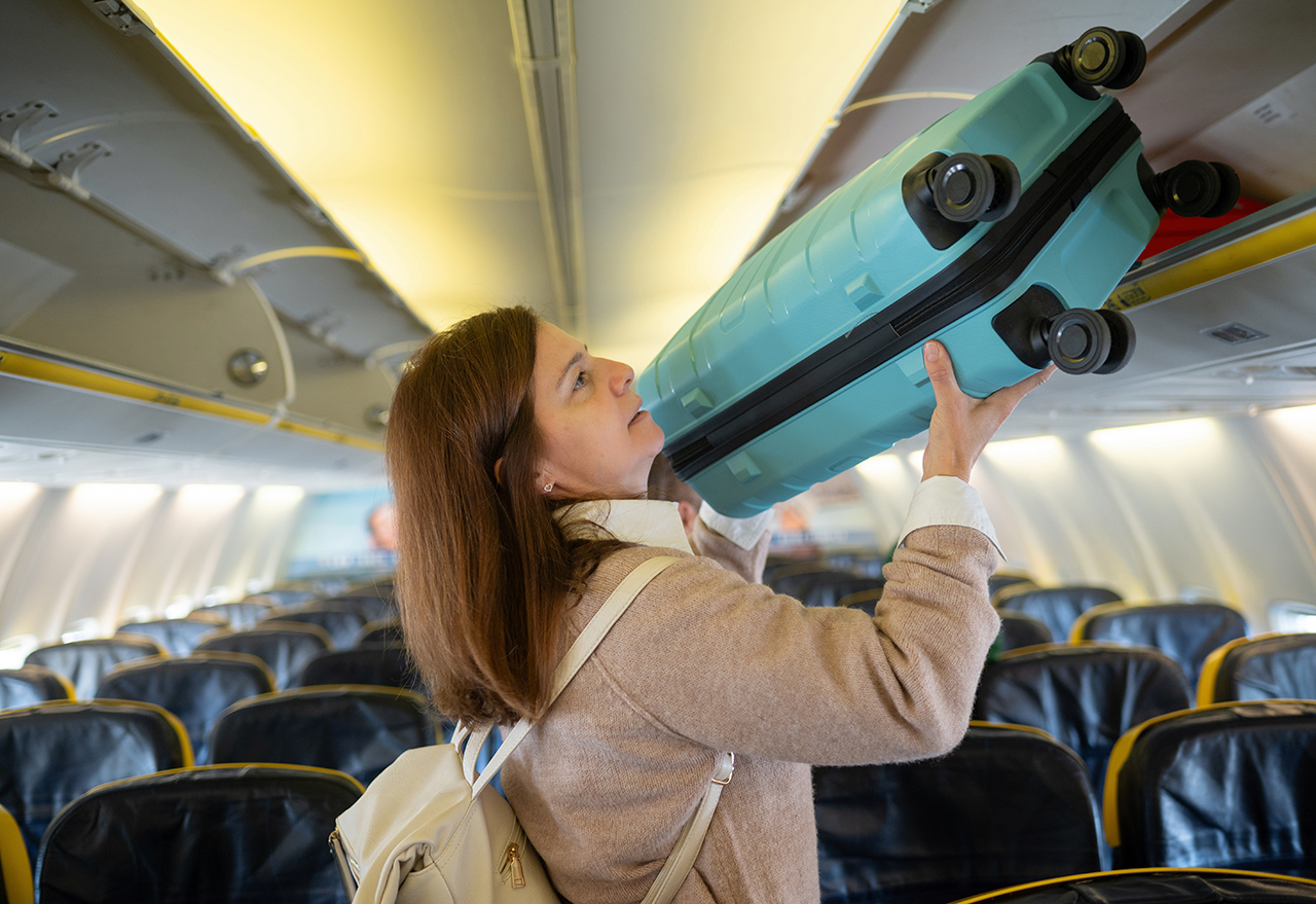 Side view of young female in casual outfit with backpack lifting luggage into compartment of airplane cabin while standing by leather chairs inside airplane